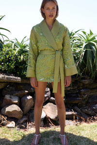 Woman wearing a green patterned outfit standing outdoors with plants and rocks in the background