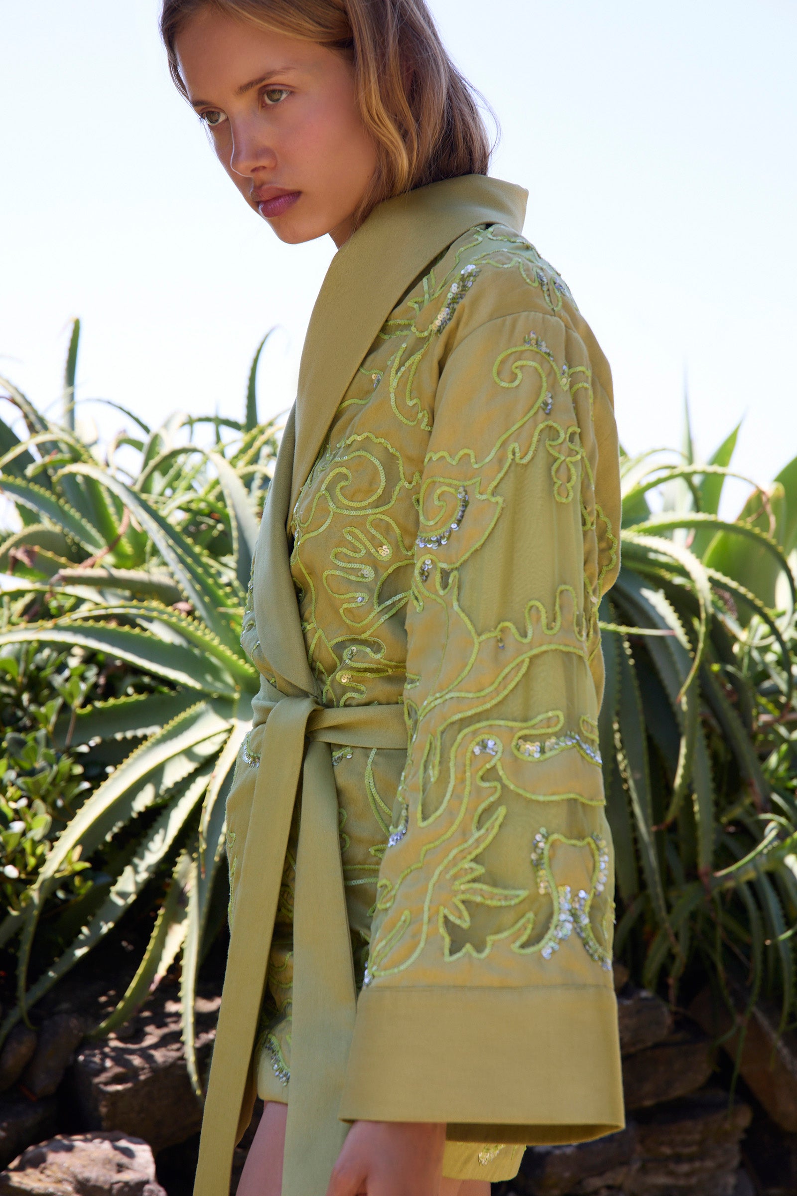 Woman wearing a light green embroidered robe in a natural setting with plants.